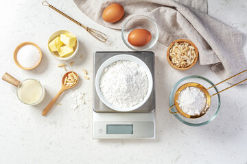 Flour in white bowl measuring on digital scale with baking ingredients and utensil on marble...