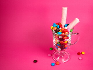 cocktail of chocolate multi-colored dragees and waffle pink tubes in a glass cup on a stem on pink background. non alcoholic cocktail. chikdhood mood. sweet party