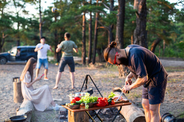 Naklejka na ściany i meble A man with a beard and black glasses sharpens a knife in nature while cooking. Outdoor recreation in summer. Bright and fresh vegetables on the table.