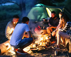 A company of friends fry sausages on a fire against the backdrop of a tent at night. Camping.