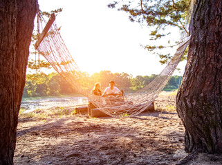 Against the background of a hammock, a guy and a girl are sitting on the sandy bank of the river in the forest. Outdoor recreation in summer, picnic. Copy space for text