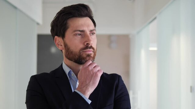 Handsome businessman in a suit looks to the side, holding his chin in a thoughtful gaze. His body language suggests that he is deep in thought, contemplating a decision or considering his next move.