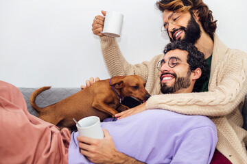 happy couple of gay men with hot drink in their mugs relax on the sofa playing with their little...