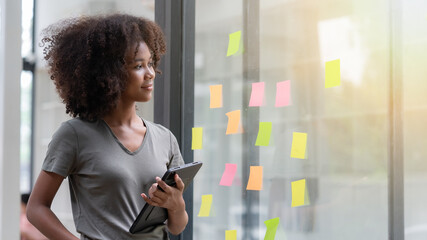 Young smiley attractive, businesswoman using sticky notes on a glass wall to write a strategic...