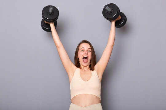 Image of beautiful happy smiling woman wearing sportswear training her biceps with weights isolated on gray background, raised arms up with heavy dumbbells, screaming with happiness.
