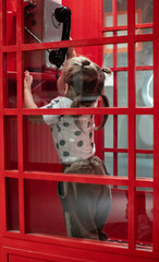A child girl hangs up in a carousel telephone booth. In a retro street phone booth, a little girl is trying to hang up after a phone call. Concept red traditional telephone booth and child.