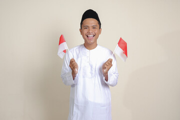Portrait of attractive Asian muslim man in white shirt with skullcap holding indonesia flag while raising his fist, celebrating success. Isolated image on gray background