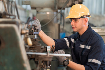 Male factory worker working at work maintenance machine in industrial factory while wearing safety uniform, glasses and hard hat. Caucasian male technician and heavy steel lathe machine in workshop