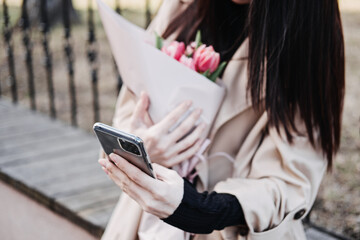 Spring outdoor portrait of happy woman with tulips bouquet, smartphone and coffee to go on spring city street. Happy young woman holding bouquet of pink flowers