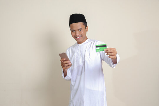 Portrait of attractive Asian muslim man in white shirt with skullcap holding a mobile phone and presenting credit card. Isolated image on gray background