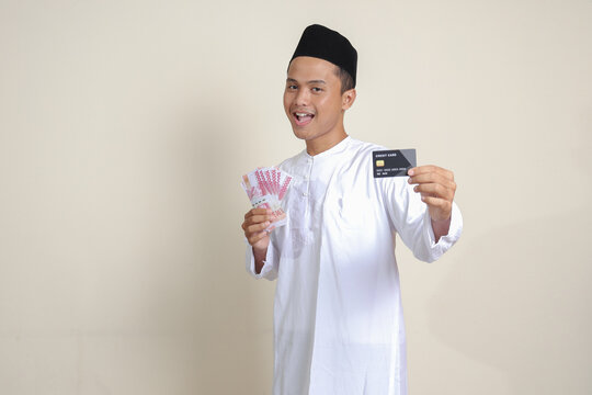 Portrait of attractive Asian muslim man in white shirt with skullcap holding one hundred thousand rupiah and presenting credit card. Isolated image on gray background
