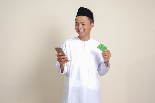 Portrait of attractive Asian muslim man in white shirt with skullcap holding a mobile phone and presenting credit card. Isolated image on gray background