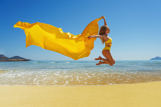 Happy young woman with a yellow light one Cloth jumping at sand beach. Relaxing, fun, and enjoy holiday at tropical paradise beach with blue sky. Girl in summer vacation.