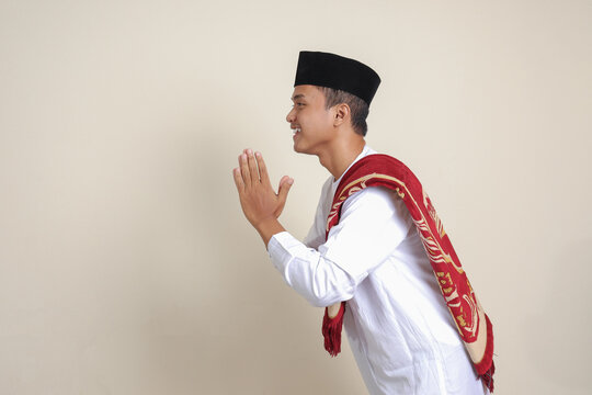 Portrait of attractive Asian muslim man in white shirt with skullcap showing apologize and welcome hand gesture. Isolated image on gray background
