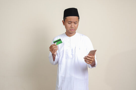 Portrait of attractive Asian muslim man in white shirt with skullcap feeling sad and disappointed, looking at his mobile phone while holding credit card. Isolated image on gray background