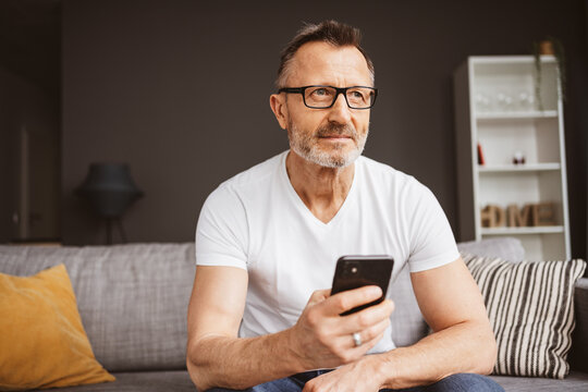 An adult man sits on a sofa in his living room, using glasses and a smartphone to stay connected.