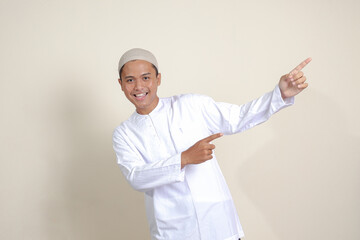 Portrait of attractive Asian muslim man in white shirt with skullcap showing product and pointing with his hand and finger to the side. Advertising concept. Isolated image on gray background