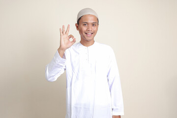 Portrait of attractive Asian muslim man in white shirt with skullcap showing ok hand gesture and smiling looking at camera. Advertising concept. Isolated image on gray background