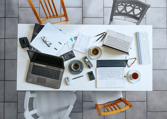 Top view of a small white meeting table with laptops, coffee, calculator, papers, ring binder and four casual chairs on a gray tiled floor business, high angle view form above