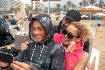 Portrait of beautiful caucasian family sitting together on the beach, mother, father and son looking at smartphone smiling