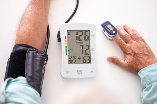 Old senior man using medical device to measure blood pressure - elderly man suffering from hypertension sitting at home table taking care of his health - Powered by Adobe