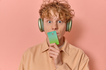 Studio shot of young surprised shocked man gen z with curly red hair enjoys music with headphones,...