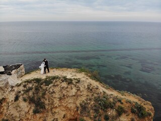 Aerial view wedding day on edge of cliff above sea, newlyweds stand with their backs on seascape background