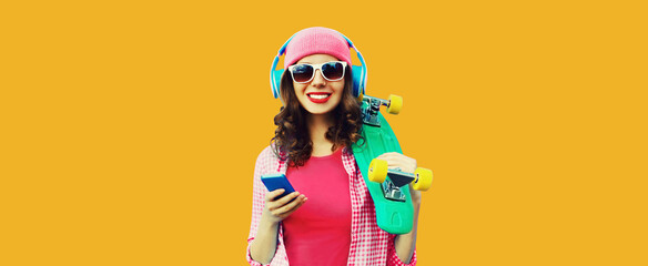 Summer colorful portrait of happy smiling young woman listening to music in headphones with smartphone and skateboard on orange background