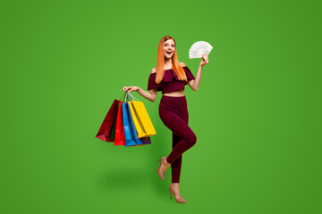 Full length size body portrait of red straight-haired beautiful young girl, wearing suit, pants, top blouse, holding shop bags and fan of dollars cash. Isolated over bright vivid yellow background