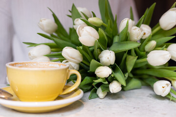 Fototapeta na wymiar Yellow ceramic mug with cappuccino is on table in cafe, next to it lies luxurious bouquet of tulips. Floral surprises. 