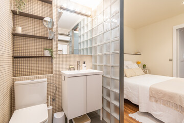 Open compact bathroom adjoining the master bedroom with glass bricks with a beige tiled sink, toilet and shelves. Concept of small but thoughtful modern apartment
