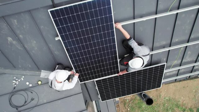 Aerial top view of technicians installing photovoltaic solar panels on roof of house. Men engineers in helmets building solar module system with help of hex key. Concept of renewable energy.