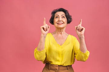 Portrait of attractive cheerful beautiful senior mature aged woman demonstrating copy space isolated over pink background, pointing fingers up.