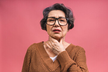 An elderly aged mature senior woman wrinkled her face and holds her hand near the throat, sore throat health problems isolated over pink background. - Powered by Adobe