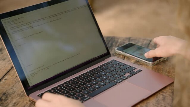 Female freelancer with computer checking statistics charts on smartphone. Woman working remotely on computer checks diagrams on phone. Female analyst studying different graphs. Remote work concept