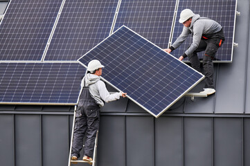 Roofers installing solar panel system on roof of house. Men workers in helmets carrying...