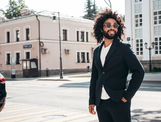 Handsome smiling hipster  model.Sexy unshaven Arabian man dressed in summer suit jacket clothes. Fashion male with long curly hairstyle posing in street at sunset. Cheerful and happy. In sunglasses