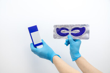 Close up unrecognizable hand in blue gloves hold and present gel and cream facial eye mask on white studio background.
