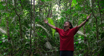 Young tourist man wearing red t-shirt raises his arms and looks up to the sky in the middle of an amazonian forest
