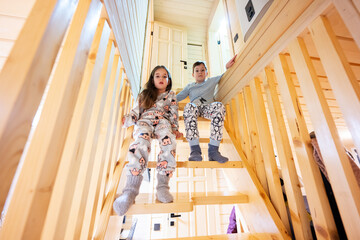 Brother with sister in pajama sit on stairs in cozy wooden tiny cabin house. Life in countryside.