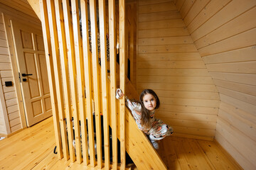 Little cute girl in pajama sit on stairs in cozy wooden tiny cabin house. Life in countryside.