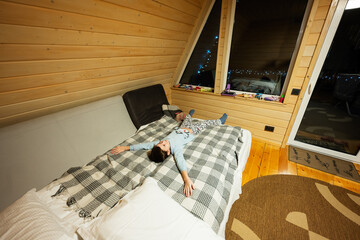 Boy lies and rests on the bed in cozy wooden tiny cabin house. Life in countryside.