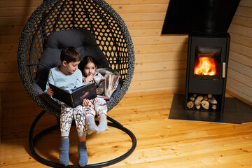 Brother with sister reading books in comfortable hanging chair against fireplace in cozy wooden...