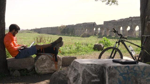 Cyclist with bike sitting on a rock under a tree writing on laptop in front of ancient roman aqueduct. Young attractive athletic man in parco degli acquedotti in Rome