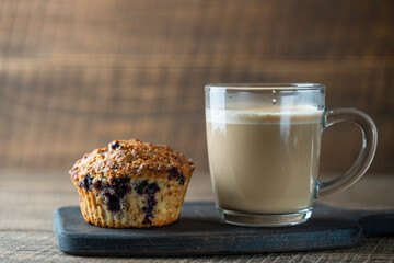 Delicious muffin with blueberries and cappuccino glass cup on a wooden table, closeup. Fresh cupcake and coffee for breakfast