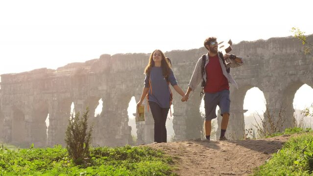 Young lovely couple backpackers tourists walking holding hands toward roman aqueduct arches in parco degli acquedotti park ruins in rome on romantic misty sunrise
