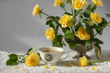 Still-life with porcelain cup of coffee, yellow garden rose flowers on pin frog in vintage vase on white lace tablecloth