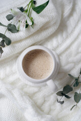 Obraz na płótnie Canvas A cup of cappuccino and flowers on a white knitted background