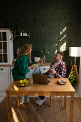 A happy couple in the kitchen sweetly talking to each other. A man with a cup of coffee in his hand sits in front of a laptop and laughs looking at his wife
