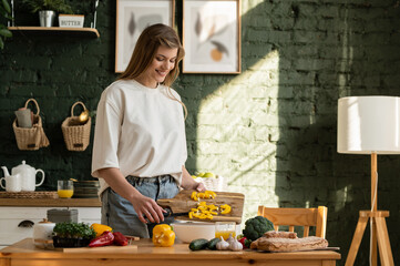 Young happy woman in the kitchen pouring sliced yellow bell pepper from a wooden cutting board into a white plate.